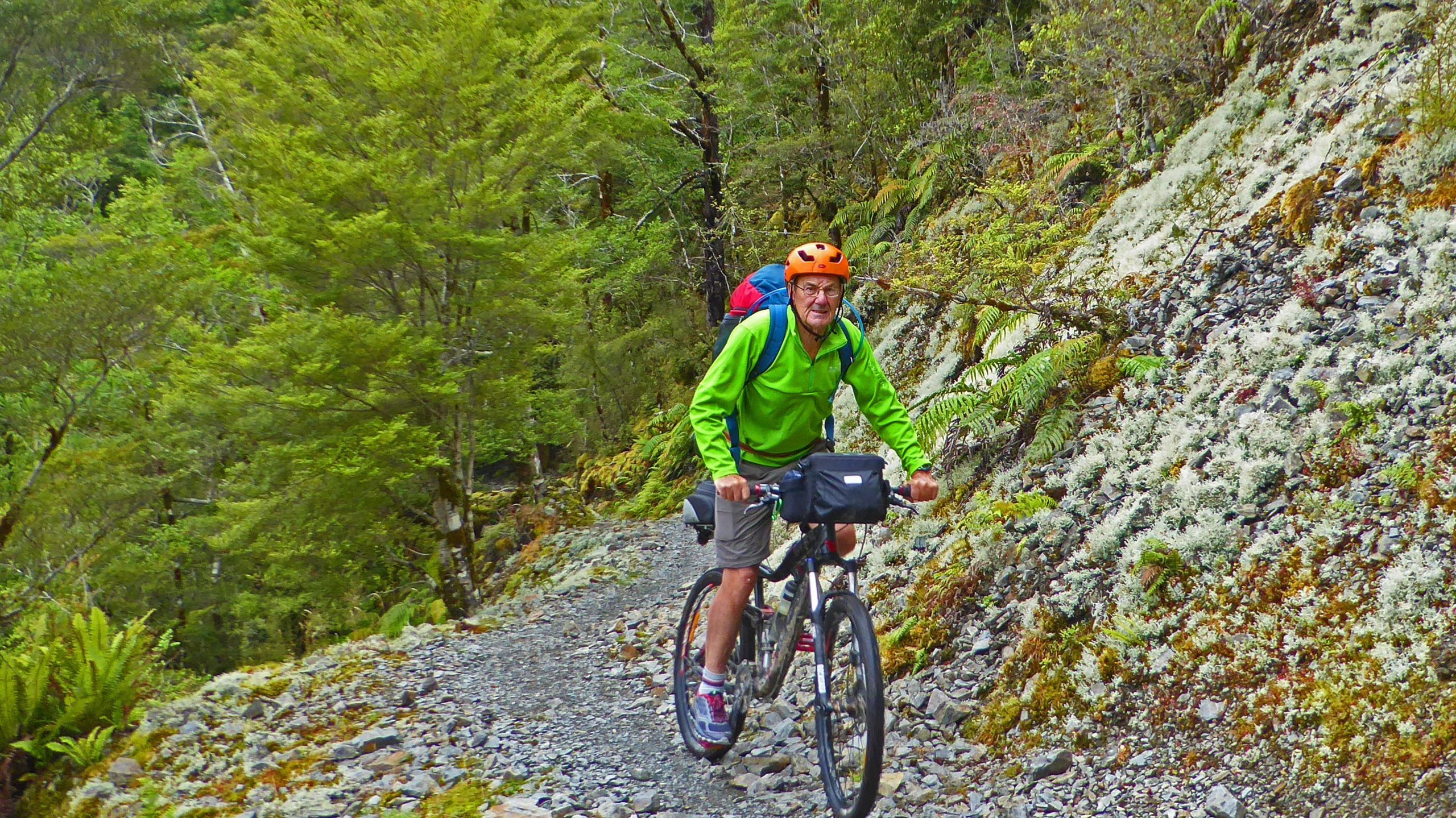 A cyclist wearing a bright green jacket and helmet rides along a rocky trail surrounded by lush greenery and trees. The cyclist has a backpack and is navigating a natural path in a forested area. The Old Ghost Road mountain bike trail.