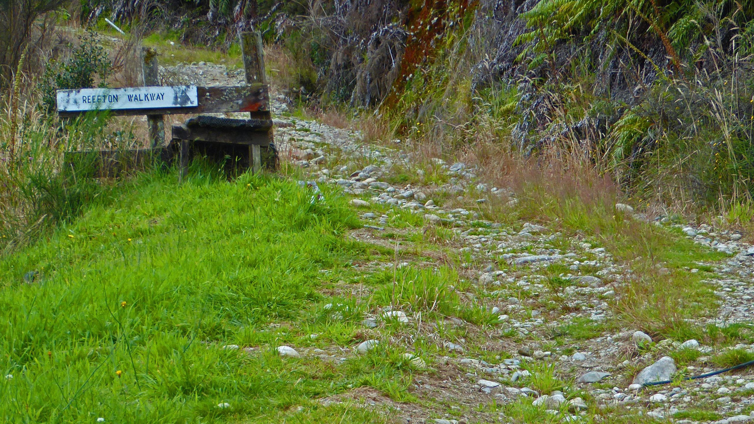 A dirt path leading past a wooden sign that reads "Reefton Walkway." The walkway is surrounded by grassy areas and sparse vegetation, with smooth stones lining the trail. The scene is set in a natural landscape, suggesting a serene outdoor environment. Reefton Walkway mountain bike trail.
