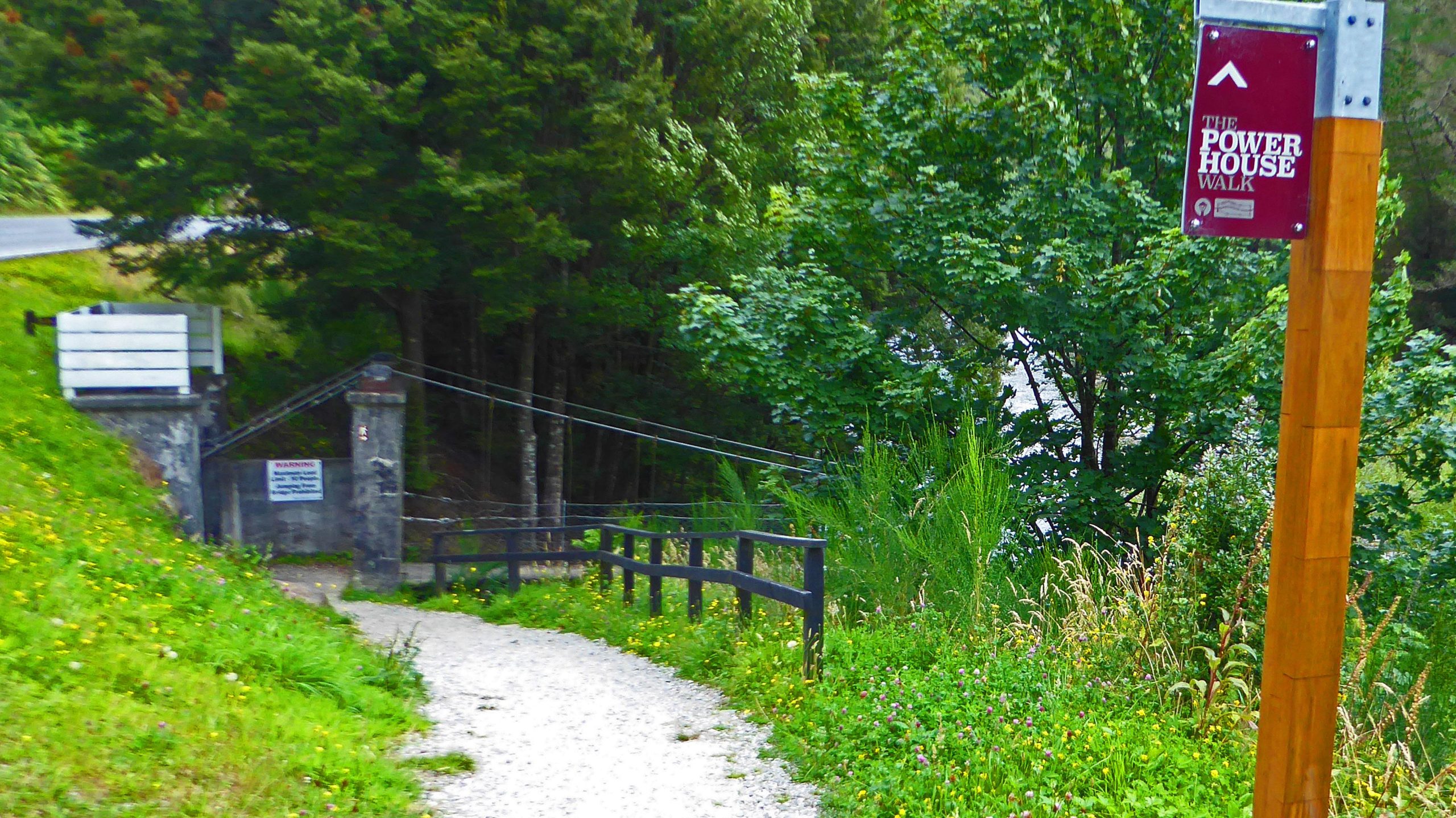 A pathway leading to a scenic area, marked by a sign for "The Power House Walk." The trail is lined with greenery, including tall grasses and flowering plants, and includes a wooden fence. In the background, there are trees and a glimpse of a river. A concrete structure with a warning sign is visible nearby. Bottled Lightening Powerhouse Loop mountain bike trail.