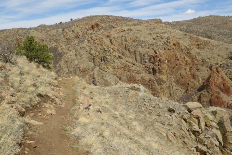 A dirt path winds along a rocky hillside, surrounded by sparse vegetation and dry grass. The landscape features rugged, multicolored rock formations and distant mountains under a clear blue sky with a few clouds.