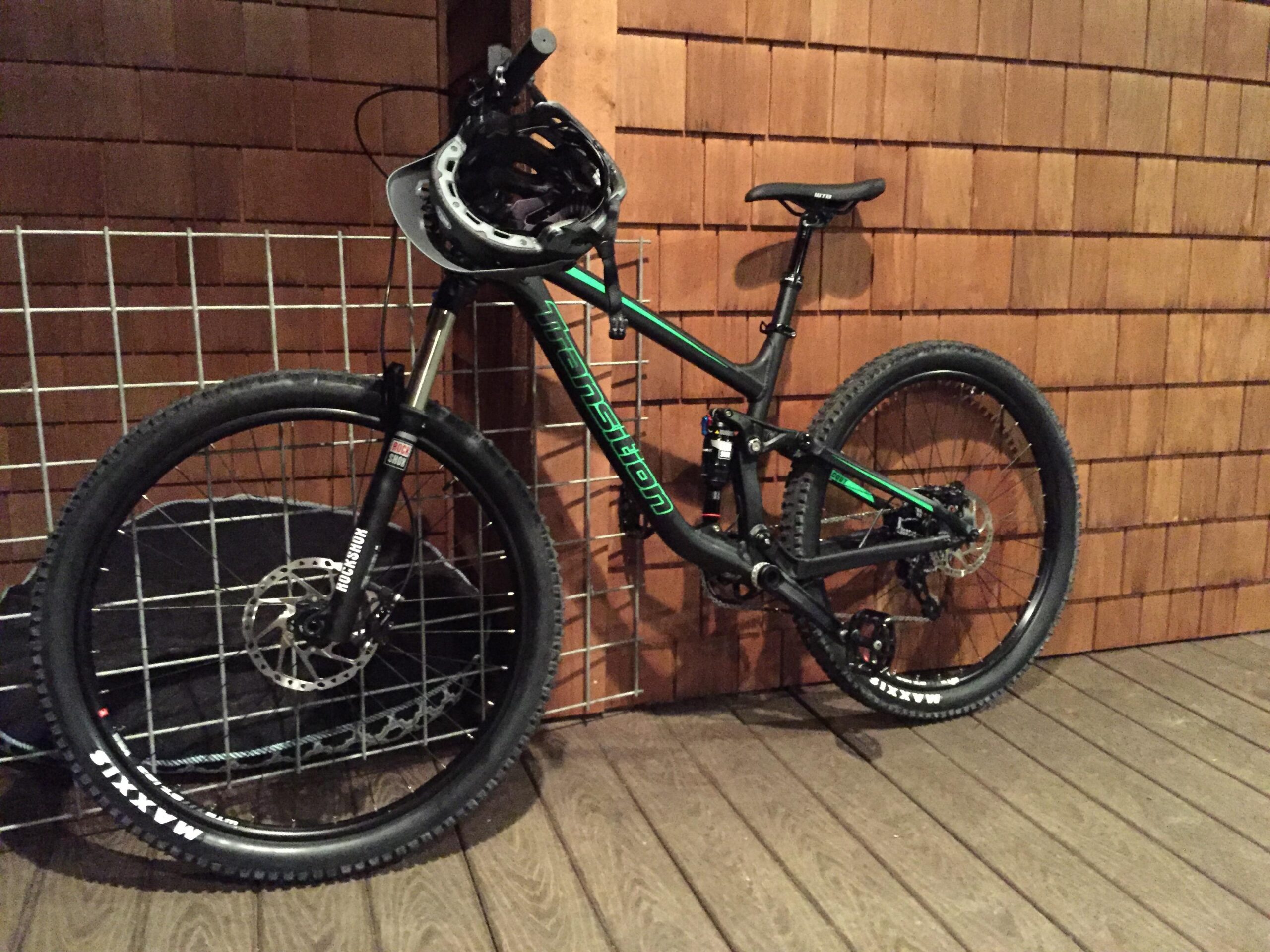 Transition Scout: A mountain bike with a black frame and green accents is leaning against a wooden wall, with a bike helmet placed on the handlebars. The bike features thick tires and a suspension fork, indicating it's designed for rugged terrain. The background includes a metal grid structure.