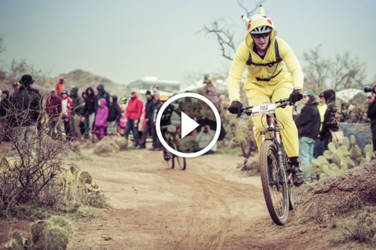A person dressed in a yellow Pikachu costume rides a mountain bike along a dirt path, surrounded by a crowd of spectators in various clothing. The scene features a rugged landscape with sparse vegetation and rocky terrain.