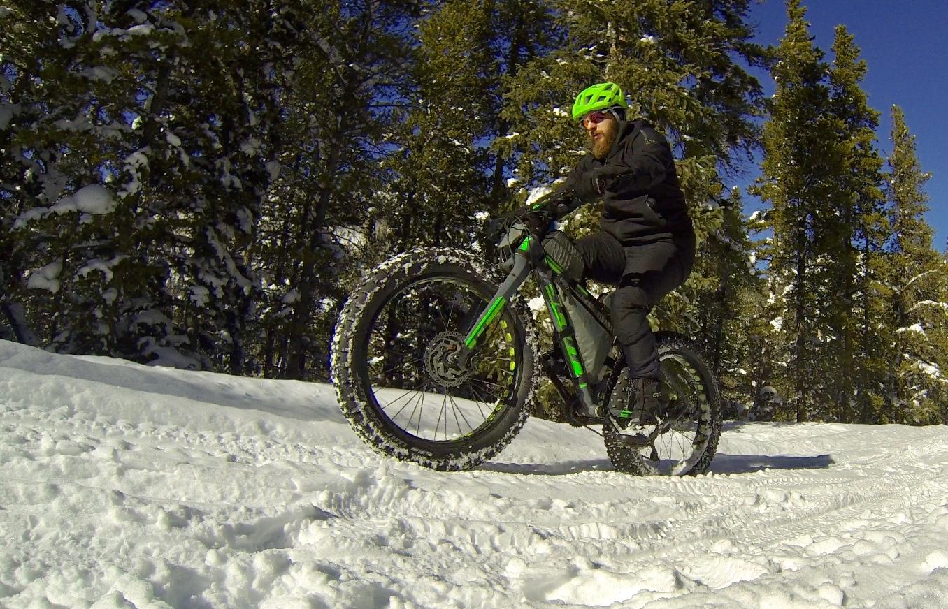 A person riding a fat tire bike on a snowy trail surrounded by trees. The cyclist is wearing a bright green helmet and black clothing, performing a wheelie as they navigate through the snow. Hancock Road mountain bike trail.