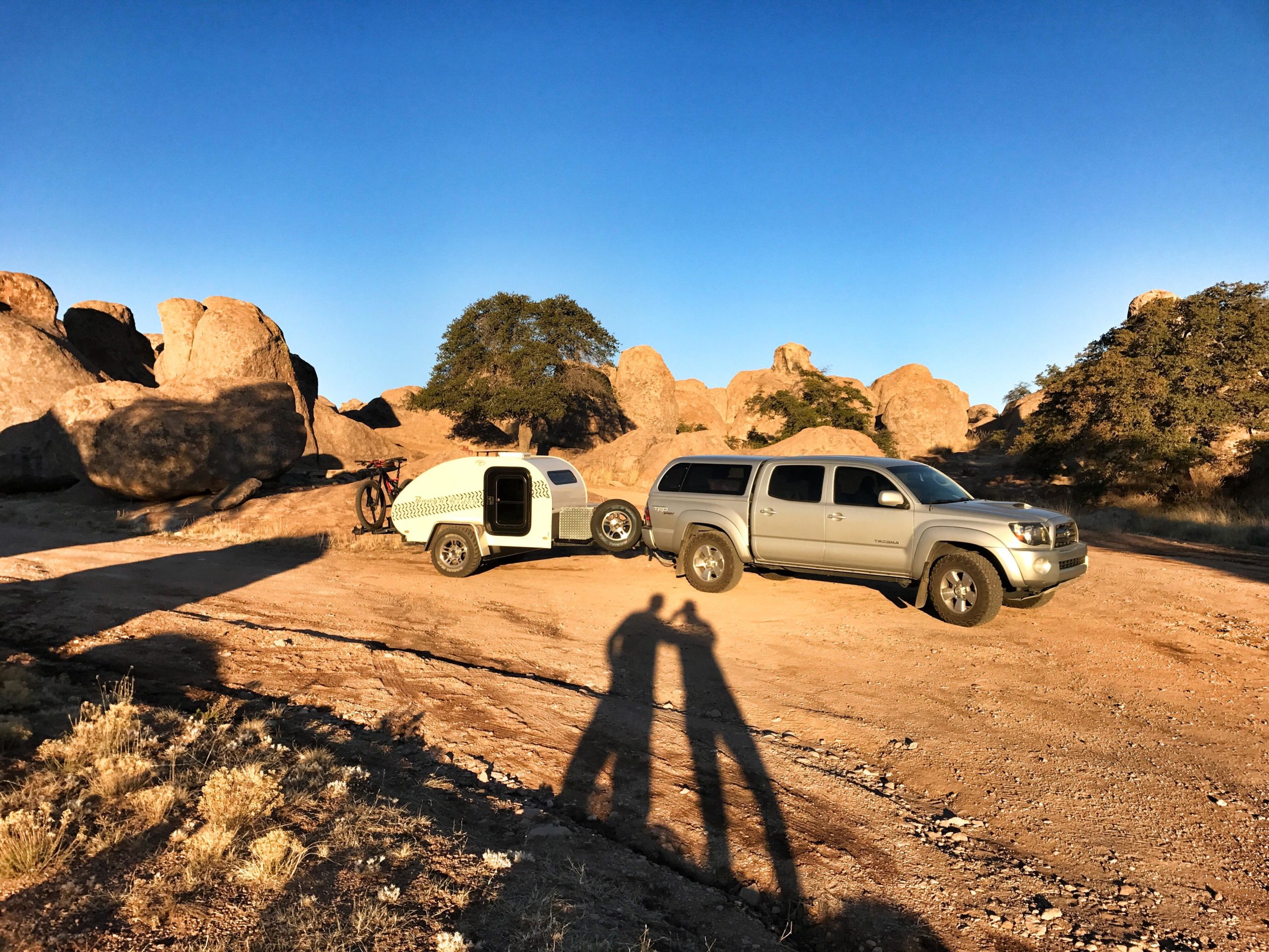 A silver pickup truck is parked next to a small white trailer on a dirt road surrounded by large rocky formations and sparse vegetation. The sun is low in the sky, casting long shadows. A mountain bike is mounted on the trailer. City Of Rocks mountain bike trail.