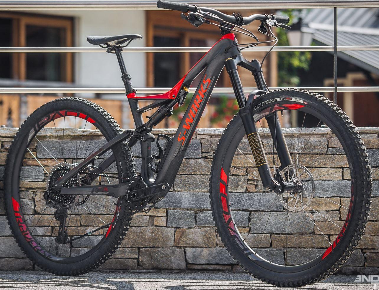 Specialized S-Works Stumpjumper FSR 29: A mountain bike with a sleek black and red design is showcased next to a stone wall. The bike features thick tires, a sturdy frame, and is equipped with an advanced suspension system. In the background, there are hints of a rustic environment with wooden structures.