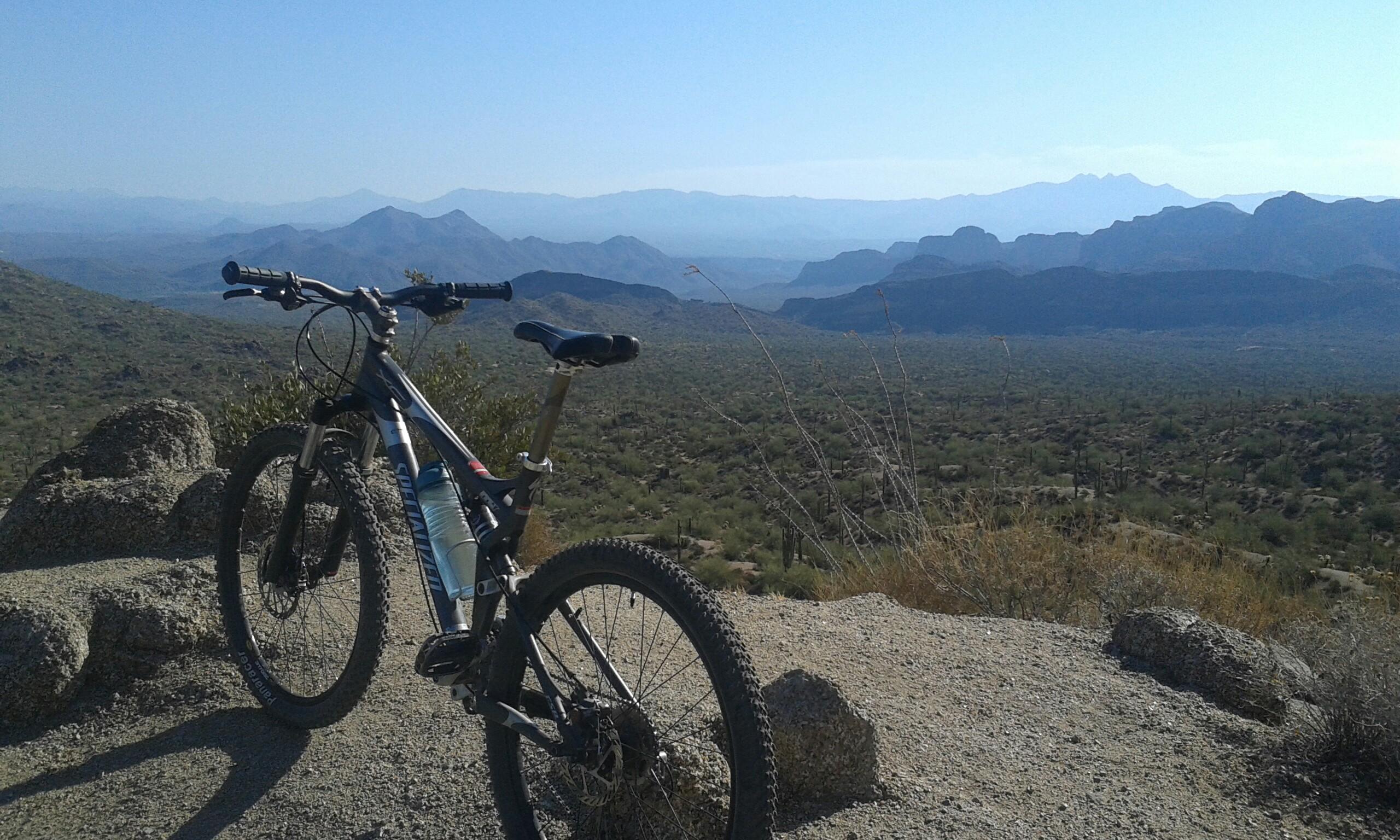 Specialized FSR: A mountain bike resting on rocky terrain, overlooking a vast desert landscape with rolling hills and distant mountains under a clear blue sky.