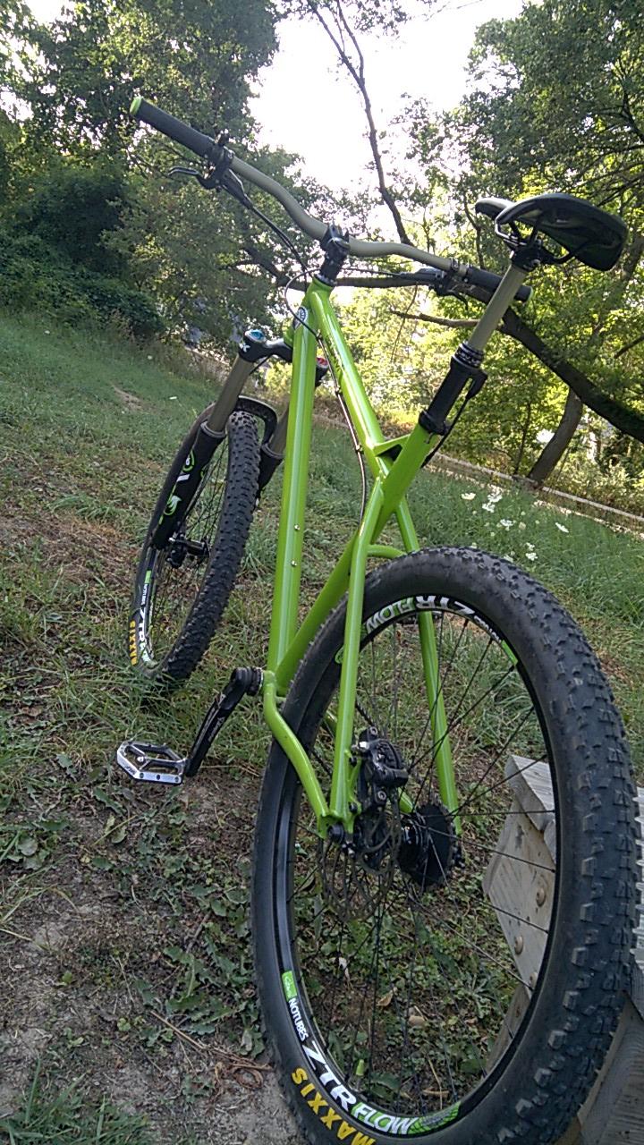 Transition TransAM: A vibrant green mountain bike positioned on grassy ground, with a backdrop of trees and a lightly shaded area. The bike is angled to showcase its frame, handlebars, and thick tires designed for rough terrain. A wooden structure is partially visible in the foreground.