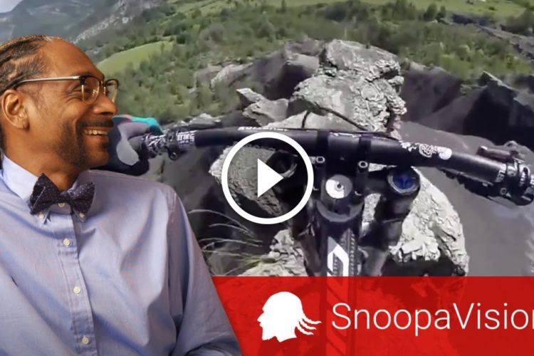 A smiling man wearing a light blue shirt and a bow tie sits beside a video featuring a mountain biking adventure, showcasing a view of rocky terrain and lush green landscapes. The video includes a play button and the text "Snoopavision" in the corner.