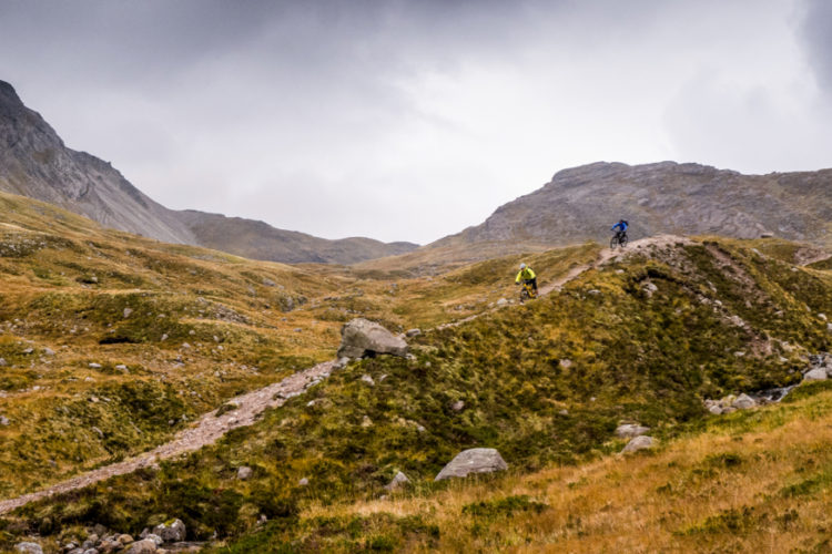 Two mountain bikers navigate a rugged, grassy terrain beneath a cloudy sky, surrounded by towering hills and rocky outcrops.