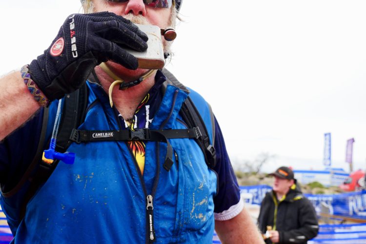 A mountain biker wearing gloves and a blue vest is drinking from a metal flask while standing in an outdoor event setting. In the background, another person is visible, and banners are set up around the area, hinting at a cycling event. The scene appears to be slightly overcast.