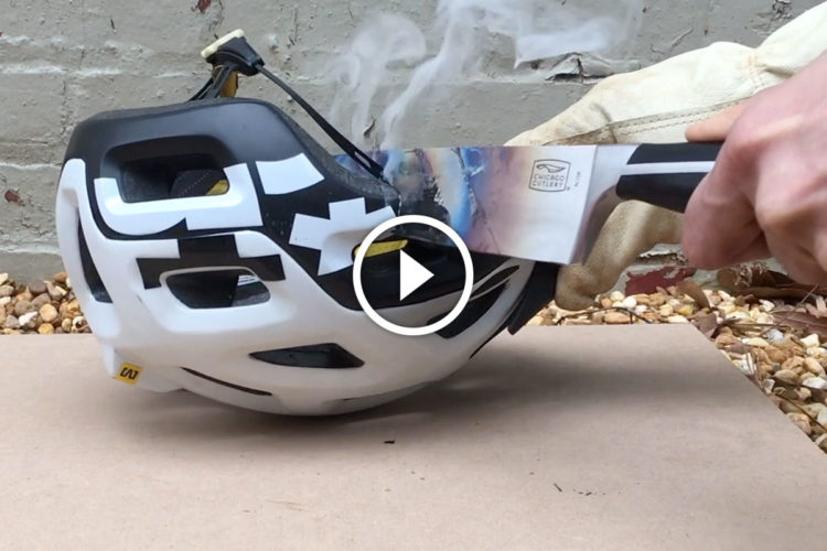 A hand is seen holding a knife, poised above a bicycle helmet that is emitting smoke. The helmet features a black and white design, with visible ventilation holes. The scene occurs on a flat surface with a gravel background, suggesting an experimental or destructive demonstration.