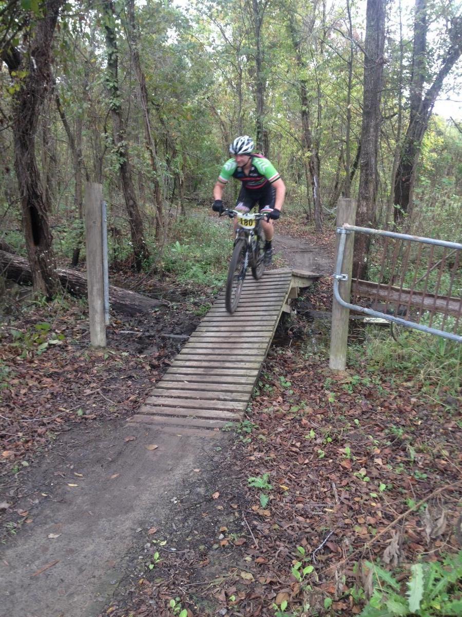 A mountain biker navigating a wooden bridge over a muddy section of a trail surrounded by trees and fallen leaves. The rider is wearing a black and green outfit and a helmet, focusing on maintaining balance while crossing the bridge. Bonnet Carre Spillway Trail mountain bike trail.