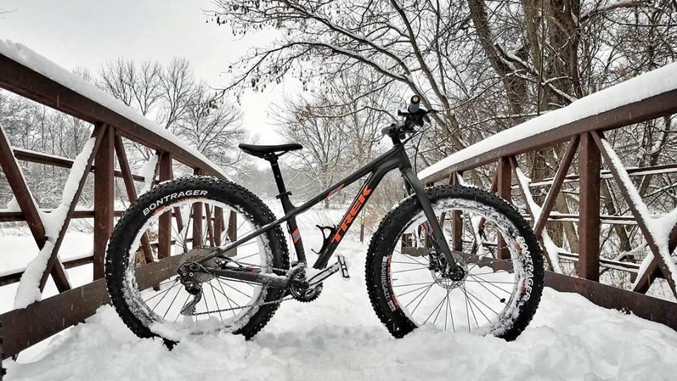 Trek farley 5: A fat tire bike resting on a snow-covered wooden bridge, surrounded by a winter landscape with bare trees. The bike features wide tires and a sleek design, showcasing its suitability for winter riding. Snow blankets the ground and the bridge, creating a serene, wintry scene.