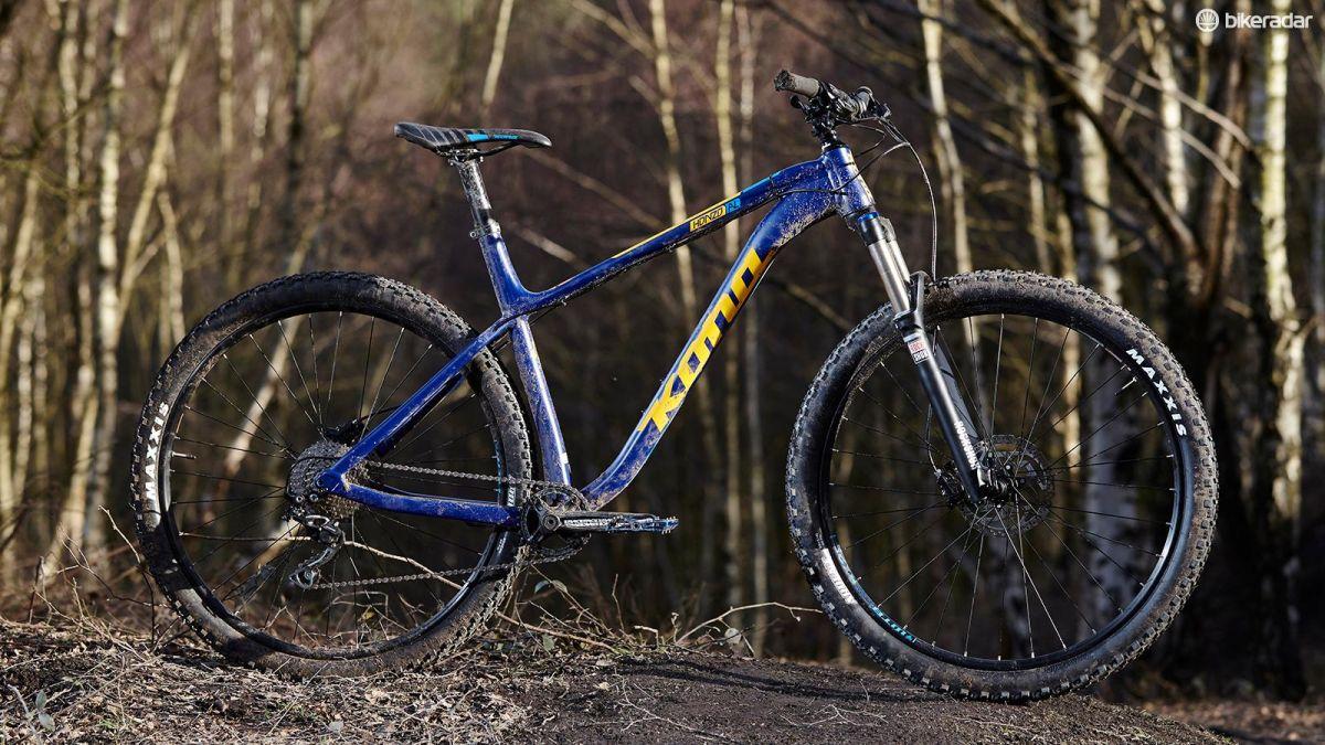 Kona Honzo: A blue and yellow mountain bike stands on a dirt trail surrounded by trees. The bike features thick tires and a sturdy frame, indicating it's designed for off-road riding. The surroundings suggest a natural, outdoor setting.