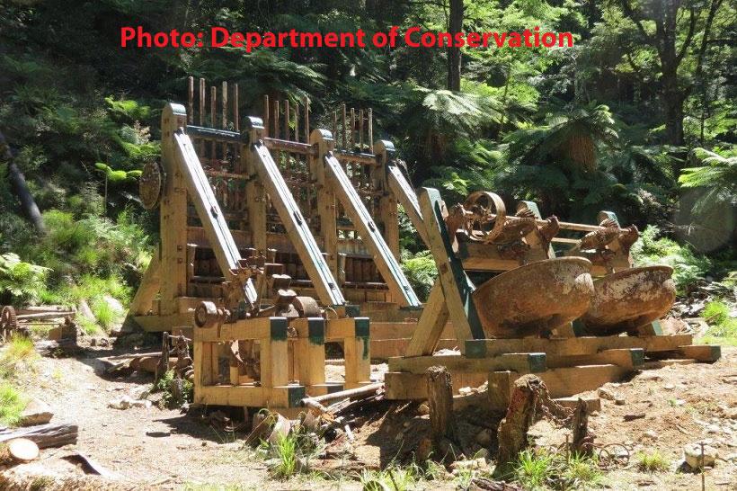 Image of historical mining equipment located in a lush forested area, showcasing a series of wooden structures with metal components. The machinery appears aged and partially overgrown by vegetation, highlighting its historical significance in the natural landscape. Montgomery Hut to Kirwans Hut mountain bike trail.