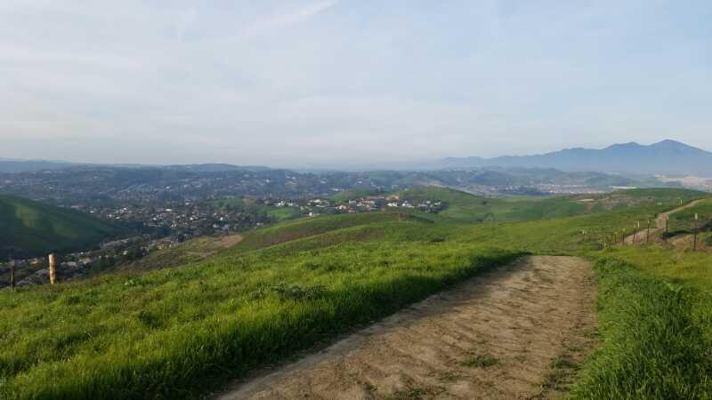 A scenic view of rolling green hills and a valley, with a dirt path leading into the foreground. In the distance, small clusters of houses are visible amidst the landscape, under a clear blue sky with some clouds. Mountains are faintly visible on the horizon. Ramblas mountain bike trail.