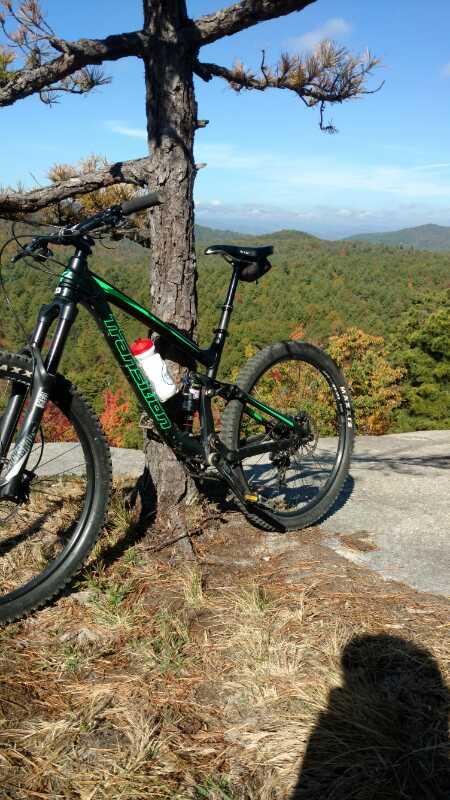 A mountain bike leaning against a tree on a rocky outcrop, with a scenic view of forested hills in the background featuring autumn foliage and a clear blue sky. A water bottle is attached to the bike frame. Cedar Rock Trail #16 mountain bike trail.