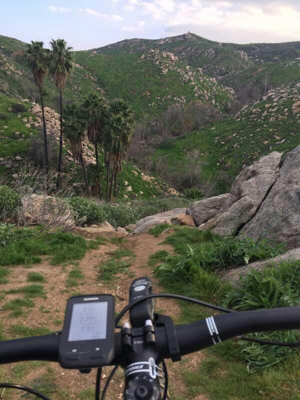 A view from the handlebars of a mountain bike on a dirt trail, surrounded by lush green hills and palm trees. In the foreground, a cycling computer displays information, while the background features rocky terrain and a scenic landscape of rolling hills. Sycamore Canyon Park mountain bike trail.