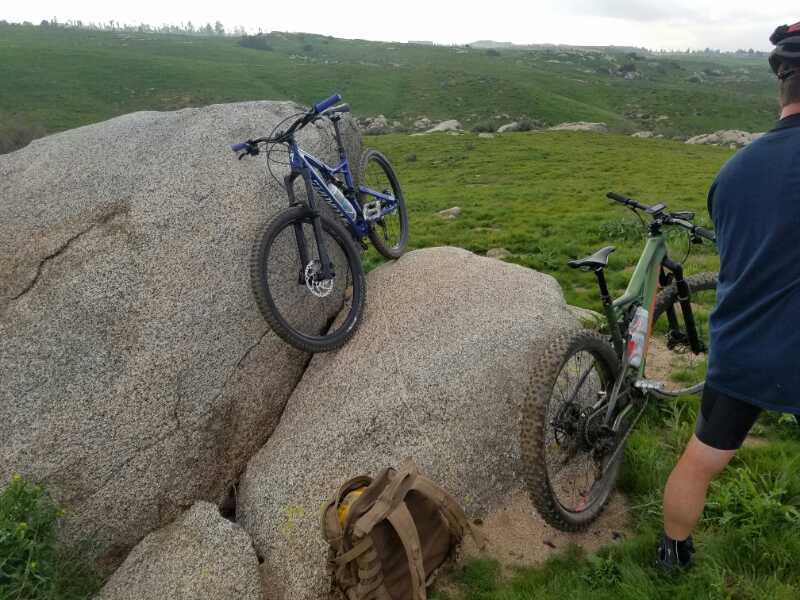 A mountain bike resting on a large rock in a green, grassy landscape, with another bike and a backpack nearby. A person in cycling gear stands partially out of frame, overlooking the scenic view. Sycamore Canyon Park mountain bike trail.