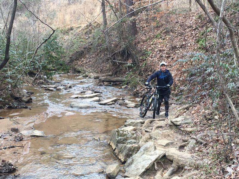 A person standing beside a rocky stream in a wooded area, holding a mountain bike. The background features trees and patches of greenery, with a slope of dried grass and earth. The setting appears to be a natural trail or outdoor recreational area. Chicopee Woods mountain bike trail.