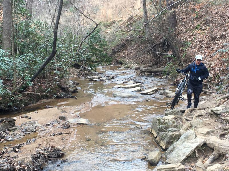 A person stands beside a rocky stream, holding a mountain bike. The surrounding area is wooded, with green shrubs and bare trees in the background. The water appears shallow, reflecting the earthy tones of the environment. Chicopee Woods mountain bike trail.