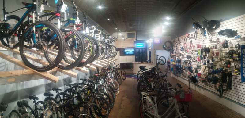 A bright bicycle shop interior featuring a variety of bikes displayed on racks and floor stands. The left side has mounted bikes on a wooden display, while the right side showcases an organized wall of cycling accessories and gear. The space is well-lit, creating an inviting atmosphere for customers.