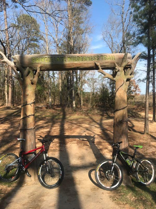 **Alt Text:** A wooden archway labeled "Bringle Lake Wilderness Trail" stands at the entrance of a dirt path surrounded by trees. Two mountain bikes are parked on either side of the archway, one red and the other black with green accents. The scene is set on a sunny day. Bringle Lake Mountain Bike Trail System mountain bike trail.