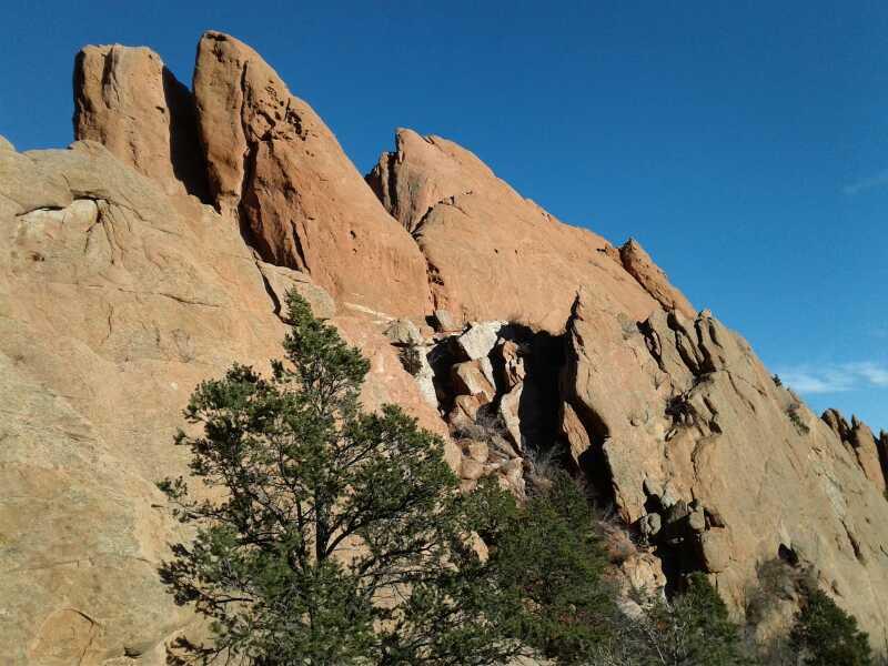 Rocky cliff formation with prominent jagged peaks and a clear blue sky in the background, featuring green bushes at the base. Garden of the Gods: Ute Trail mountain bike trail.