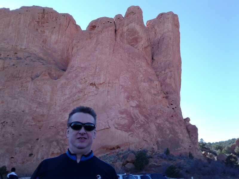A man wearing sunglasses stands in front of a large, reddish rock formation against a clear blue sky. The landscape features sparse vegetation and parked cars in the foreground. The rock formation is prominent, showcasing its towering cliffs. Garden of the Gods: Ute Trail mountain bike trail.