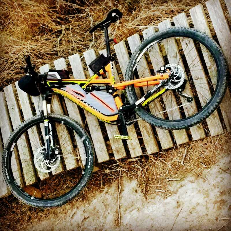 Alt text: A mountain bike with an orange and black frame is resting on wooden slats, surrounded by dry grass. The bike features thick tires and a backpack attached to the front. Oso Creek Park mountain bike trail.
