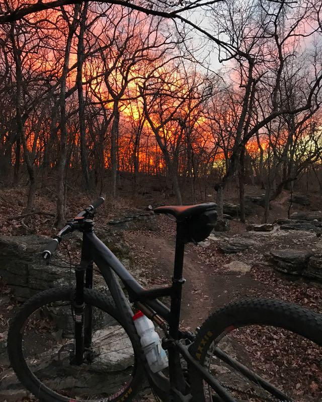 A mountain bike stands on a forest trail, with a vibrant sunset illuminating the sky in shades of orange and pink. The trees are bare, suggesting early spring or late fall, and fallen leaves cover the ground, while rocky terrain is visible in the background. Swope Park Trail mountain bike trail.