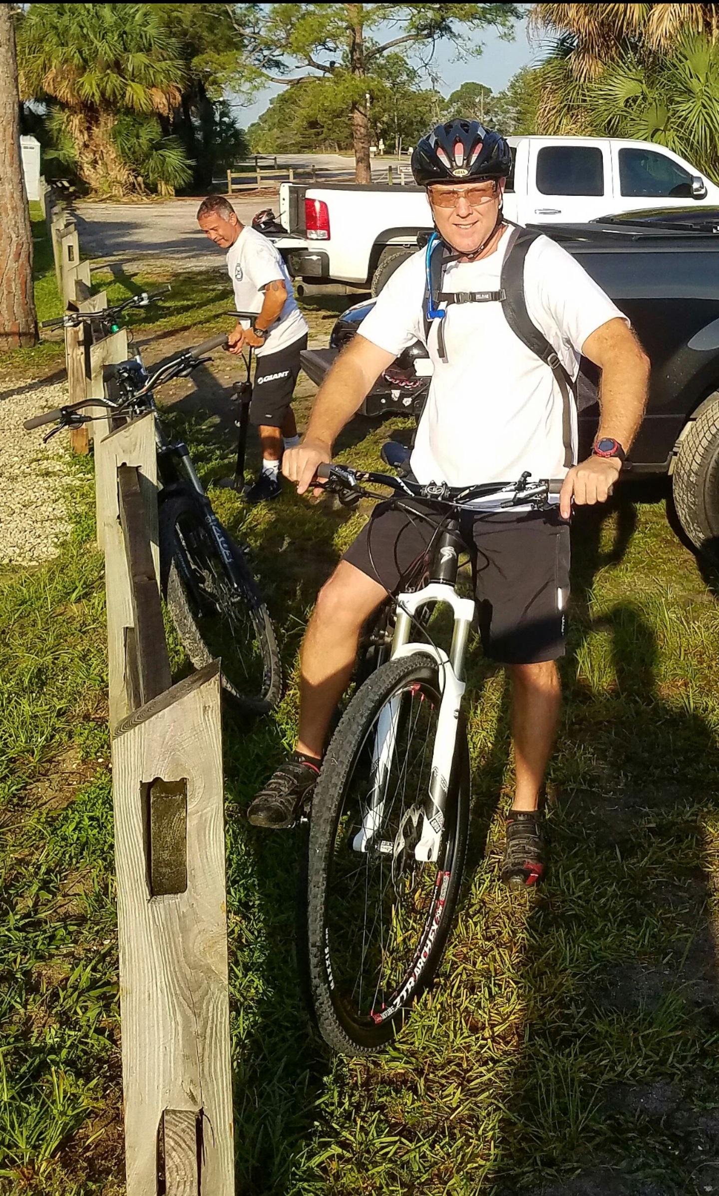 Santa Cruz Tallboy Carbon: A man wearing a helmet and a white shirt is standing next to a mountain bike, gripping the handlebars with both hands. In the background, another person is preparing a bike by a wooden fence. The scene is set in a grassy area with trees and vehicles parked nearby, indicating a recreational biking location on a sunny day.