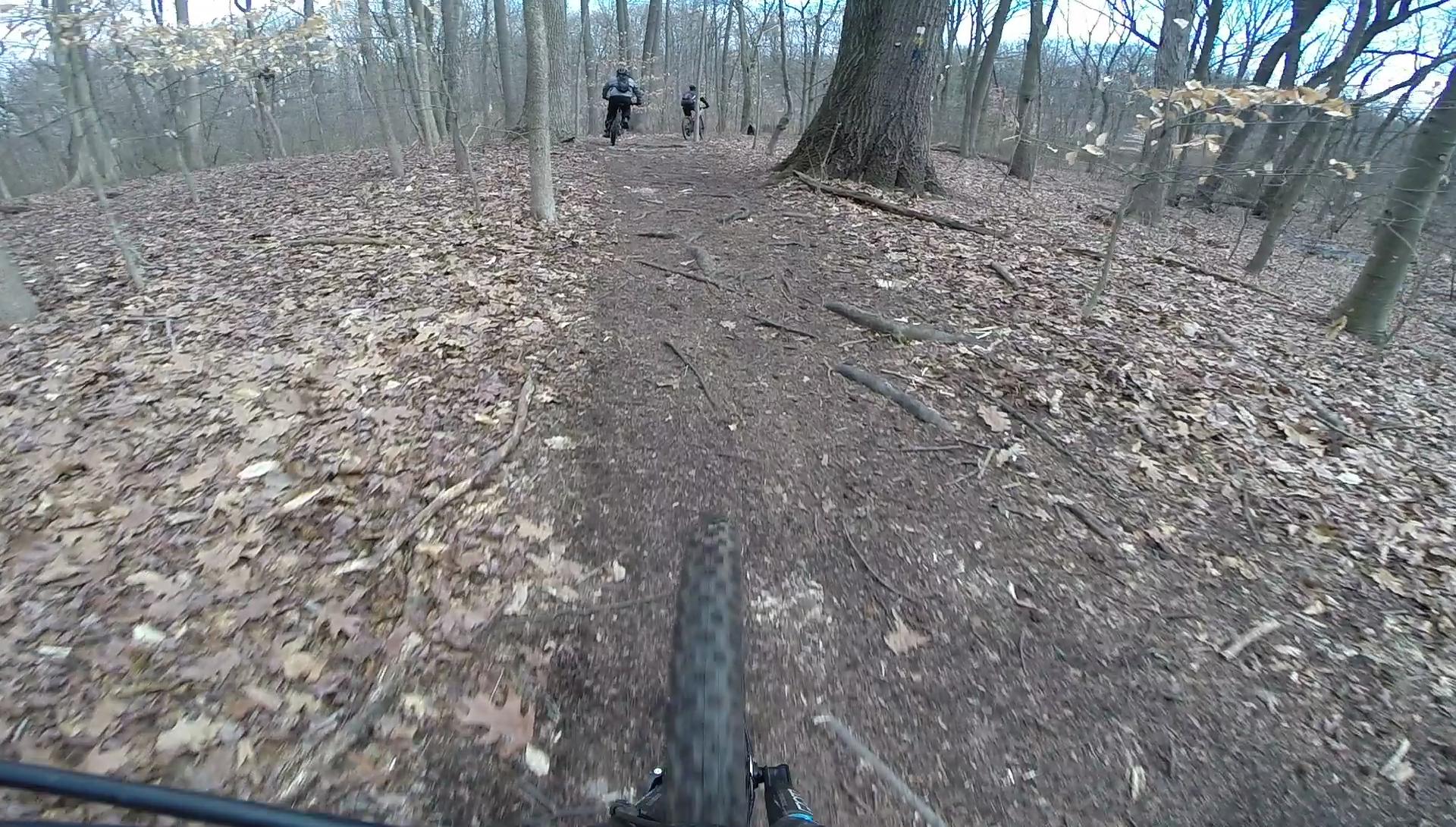 Mountain biking trail view from the front of a bike, showing a dirt path covered with leaves and roots, with two cyclists visible in the background riding along the trail in a forested area. Richmond Avenue and Forest Hill road mountain bike trail.