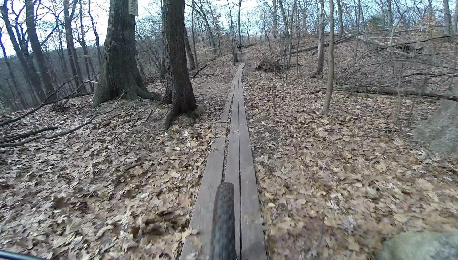 A narrow wooden pathway winding through a wooded area, surrounded by bare trees and fallen leaves, with a bicycle tire in the foreground. The scene captures a serene, natural environment ideal for outdoor biking or hiking. Richmond Avenue and Forest Hill road mountain bike trail.