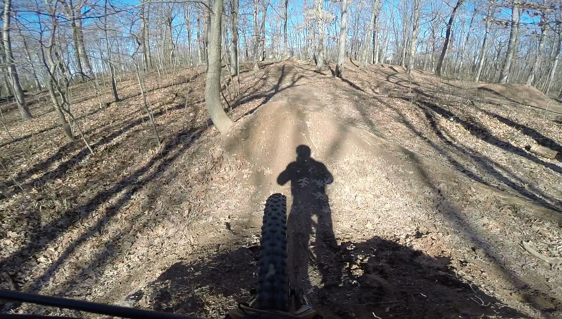 A mountain bike's front wheel and shadow are visible in the foreground, aimed towards a dirt jump in a wooded area. The scene features bare trees and a clear blue sky, with fallen leaves covering the ground. Trails and shadows create an adventurous atmosphere for mountain biking. Wolfes Pond park mountain bike trail.