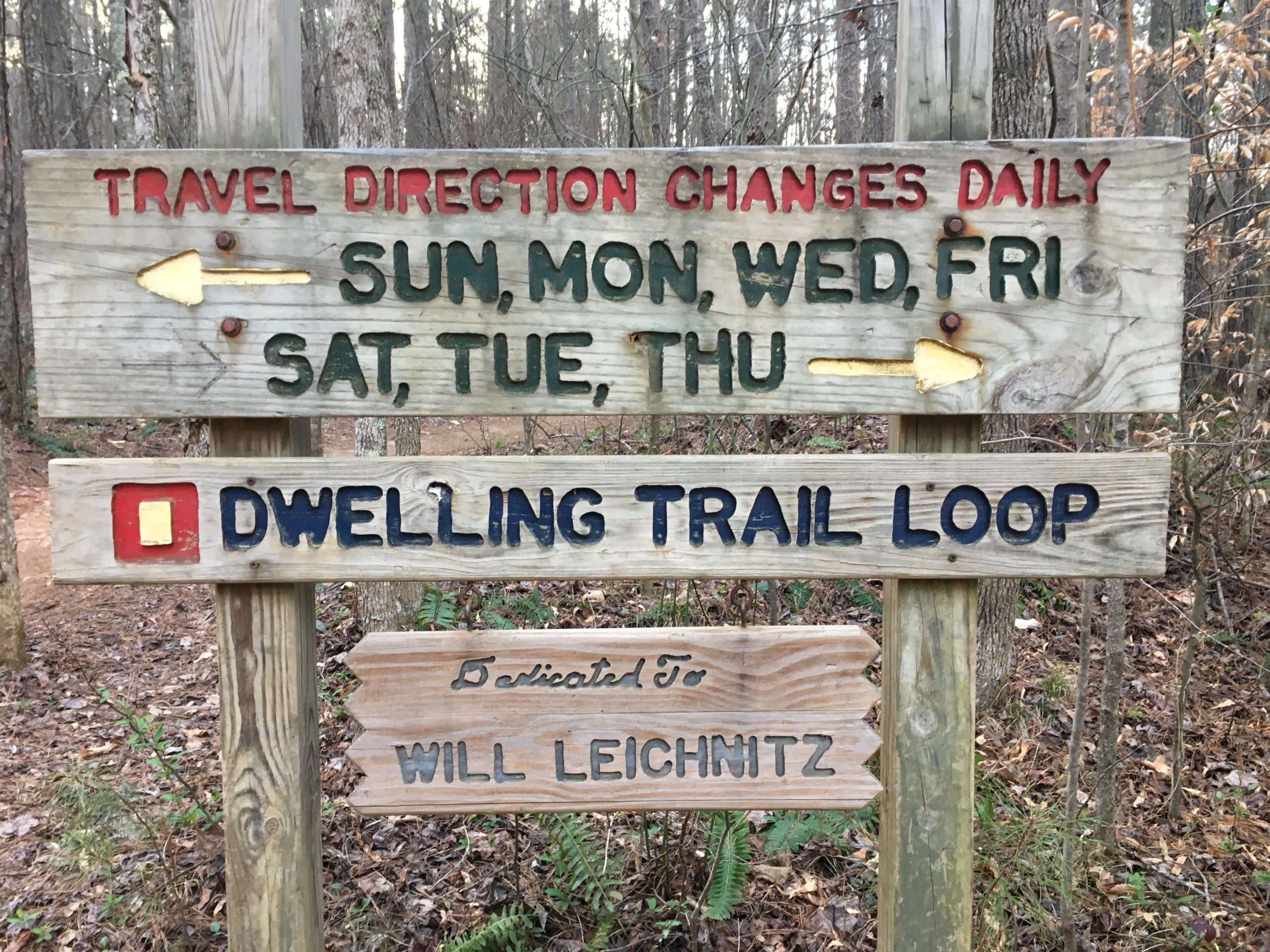 Wooden trail sign indicating daily travel direction changes, with arrows and days of the week listed. The sign features "DWELLING TRAIL LOOP" highlighted, and is dedicated to Will Leichnitz. The surrounding area includes trees and foliage, typical of a woodland environment. Dwelling Loop mountain bike trail.