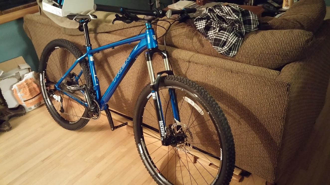 Trek Stache: A blue mountain bike resting against a brown couch, with a plaid shirt draped over the couch. The bike features thick tires, a front suspension fork, and a disc brake. The background includes a partially visible box and some other items on the floor.
