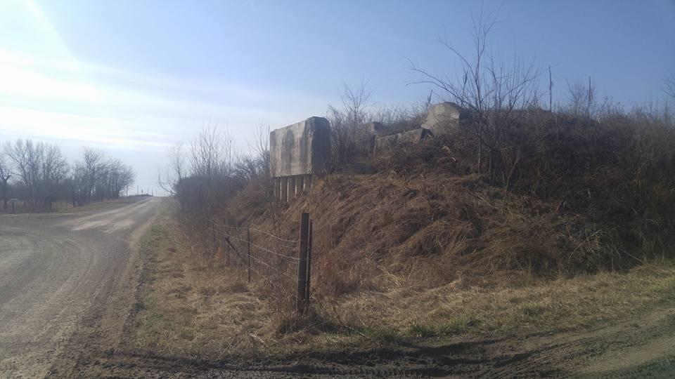 An overgrown embankment beside a dirt road, featuring a weathered structure partially hidden by tall grass and sparse trees under a clear sky. Flint Hills Nature Trail (Rail Trail) mountain bike trail.