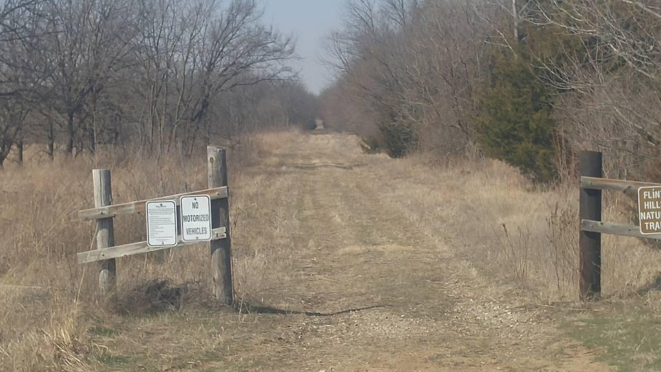 A dirt trail leads into a wooded area, flanked by dry grass and bare trees. A wooden sign at the trailhead indicates "No Motorized Vehicles," while another sign identifies the trail as "Flint Hills Nature Trail." The pathway extends straight ahead, inviting exploration. Flint Hills Nature Trail (Rail Trail) mountain bike trail.