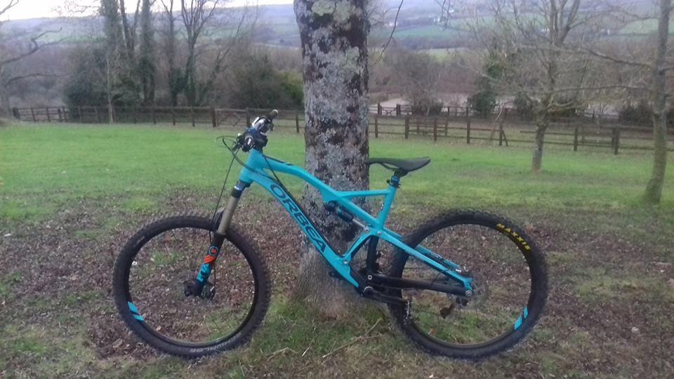 Orbea Rallon X-10: A bright blue mountain bike leaning against a tree in a grassy field, with rolling hills and a wooden fence visible in the background. The bike features thick tires and a suspension fork, indicating it's designed for off-road use.