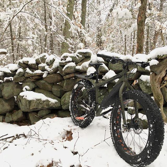 SE F@R: A fat-tire bicycle resting against a snow-covered stone wall in a forest during winter. The ground is blanketed in white snow, and the trees in the background are also dusted with snowflakes, creating a serene winter scene.