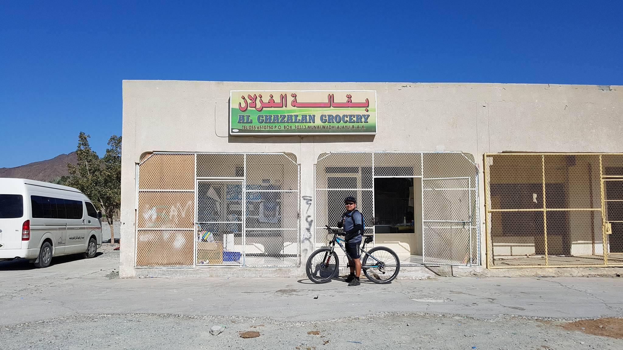 A person standing beside a bicycle in front of a grocery store named "Al Ghezlan Grocery," which features a sign in English and Arabic. The building is partially covered with metal grates, and a white van is parked nearby. The background includes a clear blue sky and mountains. Showka Trails mountain bike trail.