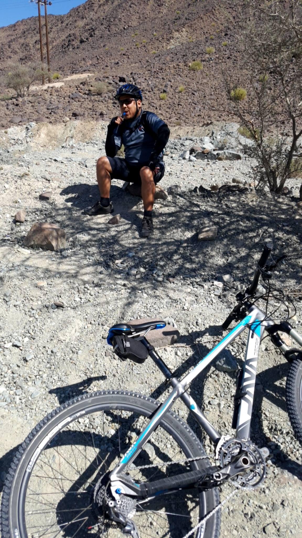 A cyclist resting on a rocky terrain, wearing a helmet and athletic clothing, while drinking from a water bottle. A mountain bike is positioned in the foreground, featuring a close-up of the wheel and frame. The background shows a dry, rugged landscape with sparse vegetation and distant hills. Showka Trails mountain bike trail.