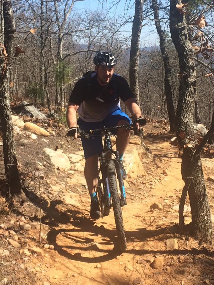 A man riding a mountain bike on a rugged trail surrounded by trees. The path is dirt and rocky, with a few loose stones scattered along the way. The rider is wearing a helmet and athletic clothing, focusing on navigating the challenging terrain. Coldwater Mountain mountain bike trail.