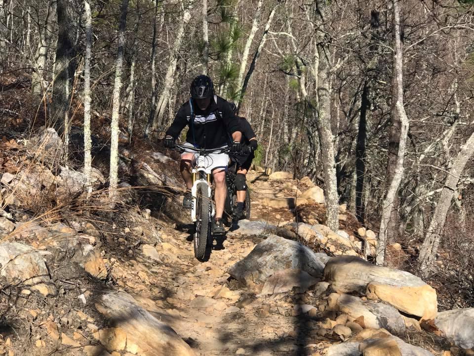 Two mountain bikers navigate a rugged trail surrounded by trees. The trail features rocky terrain and uneven ground, with one biker in the foreground focusing intently as he rides. The scene captures the challenge and excitement of outdoor biking in a natural setting. Coldwater Mountain mountain bike trail.