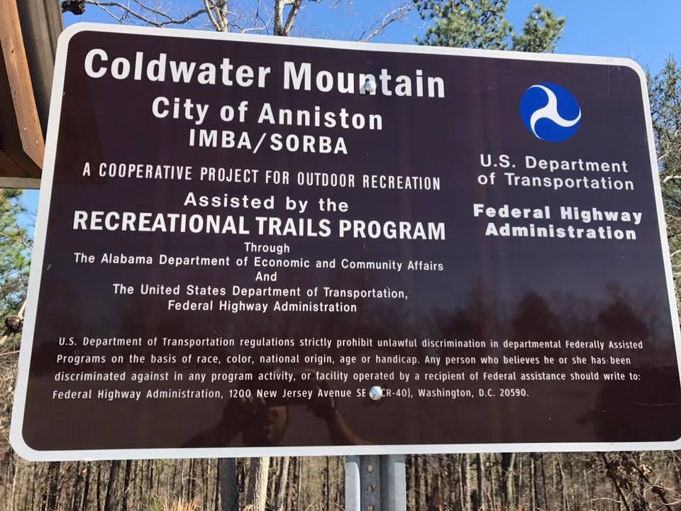 Sign for Coldwater Mountain in the City of Anniston, indicating a cooperative outdoor recreation project assisted by the Recreational Trails Program. The sign includes information about funding from the Alabama Department of Economic and Community Affairs and the U.S. Department of Transportation, emphasizing nondiscrimination in federal programs. The background features trees. Coldwater Mountain mountain bike trail.