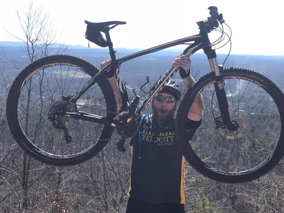 A person wearing a cycling jersey and sunglasses raises a mountain bike above their head while standing on a hillside with a scenic view of distant mountains and trees in the background. The individual has a joyful expression, celebrating their achievement in cycling. Coldwater Mountain mountain bike trail.