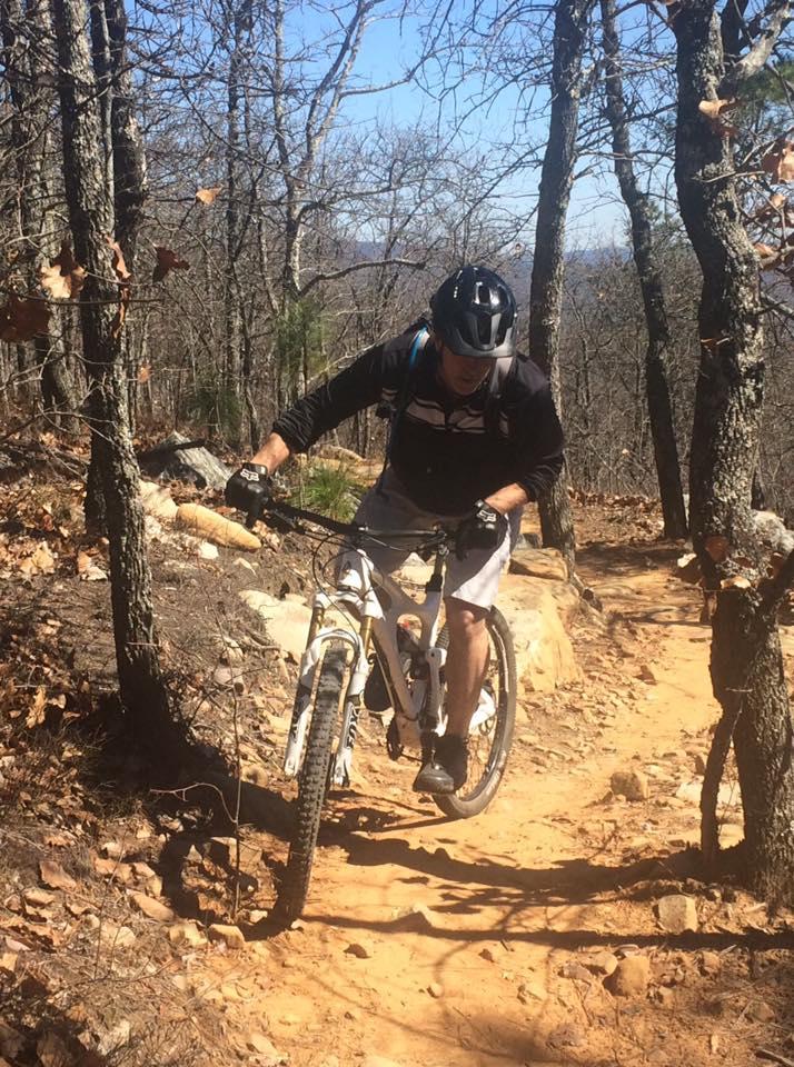 A mountain biker riding on a dirt trail surrounded by trees, with rocky terrain and sparse foliage, wearing a helmet and gloves. The cyclist is focused, navigating the winding path on a light-colored mountain bike. Coldwater Mountain mountain bike trail.