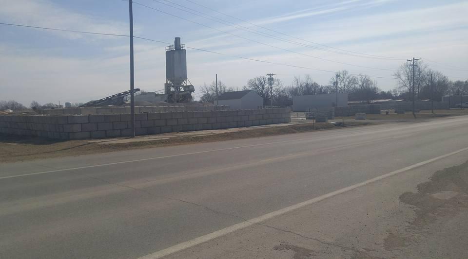 Construction site view along a rural road, featuring a concrete block wall, a concrete silo, and machinery. In the background, there are additional structures and trees. The sky is partly cloudy, indicating a clear day. Flint Hills Nature Trail (Rail Trail) mountain bike trail.