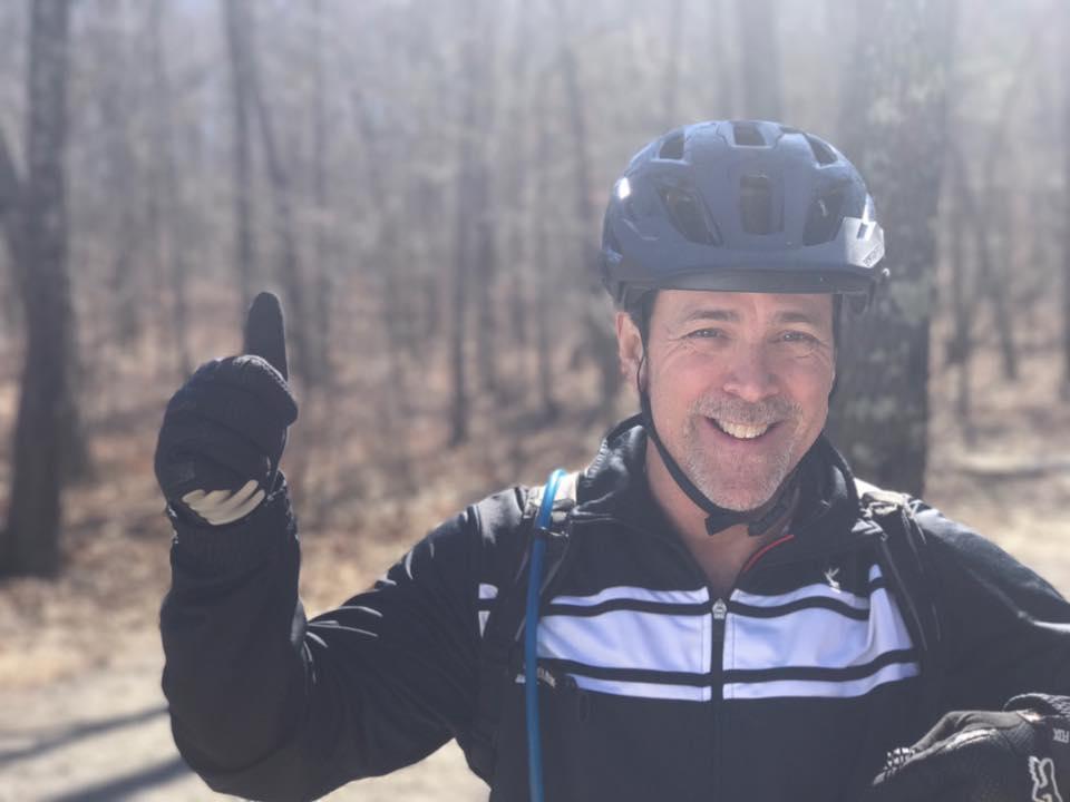 A smiling cyclist wearing a helmet and gloves gives a thumbs-up gesture while standing in a wooded area on a sunny day. Coldwater Mountain mountain bike trail.